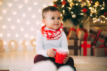 Baby with red mug sitting on the background of Christmas tree charming new year's eve and Christmas