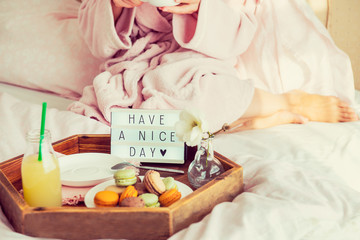 Fototapeta na wymiar Good morning concept. Breakfast in bed with Have a nice day text on lighted box, juice and macaroons on tray and blurred woman in bathrobe drinking coffee. Hospitality, care, service. Copy space.