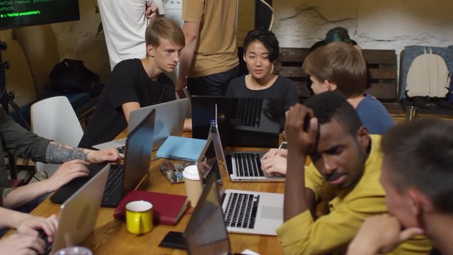 Group of young people of different ethnicities sitting at table together, typing codes on laptop computers and talking to each other