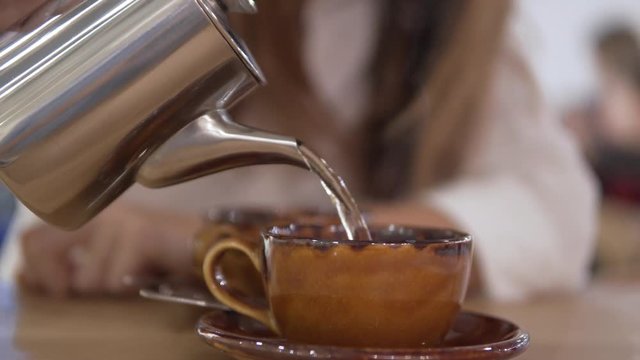 Close-up of a stylish porcelain cup and metal teapot. Female hand pours herbal green tea into an unusual cup.
