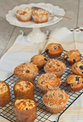 Top view, medium distance of a variety of freshly baked, homemade, muffins in paper cups, cooling on a wire cooling rack with a white pedestal display dish on wood table