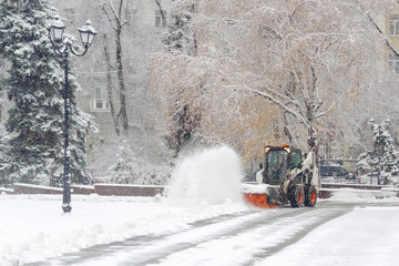 City service cleaning snow , a small tractor with a rotating brush clears a  road in the city park from the fresh fallen snow on  winter day