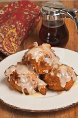 Top view, close distance of three, stacked, freshly baked, homemade, maple, glazed bacon fritters on around, white plate with a gold rim on a wood board and a print, cloth napkin and contain of syrup