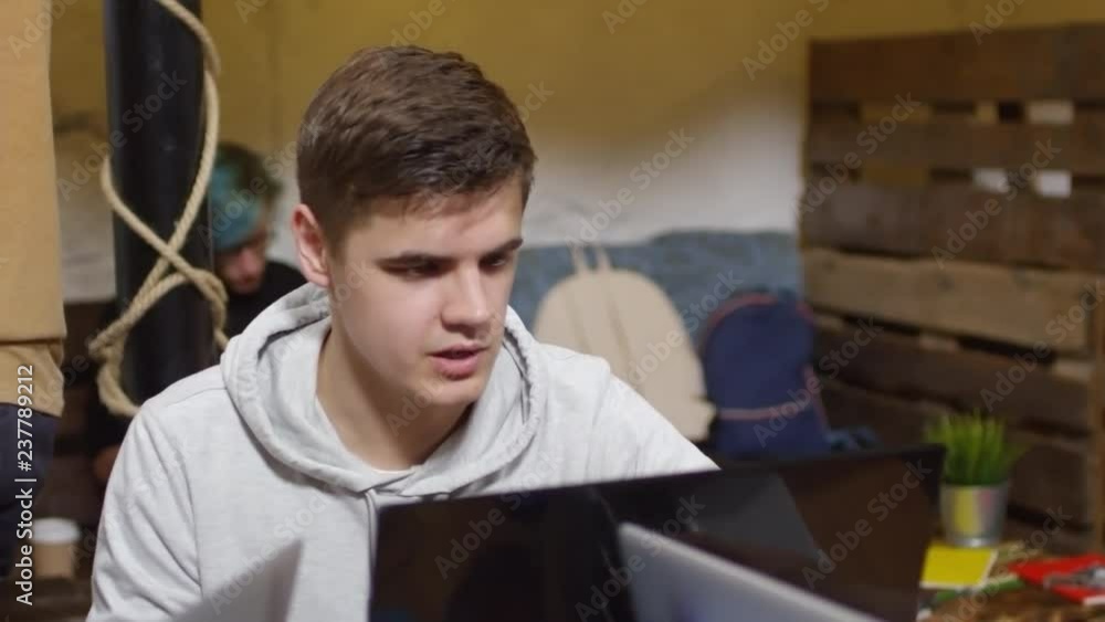 Poster medium shot of young caucasian man sitting at desk, using laptop computer and talking during program