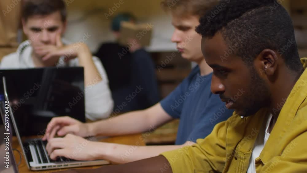 Poster medium shot of concentrated multiethnic young men sitting at desk and typing on laptop computers dur