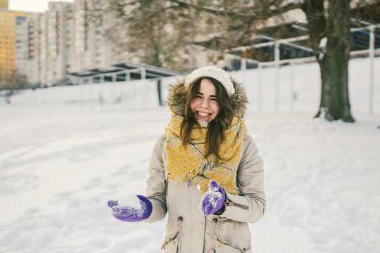 Portrait of a beautiful young Caucasian woman in a knitted hat and scarf standing on a winter background with snow smile and happiness purple gloves sculpt a snowball.