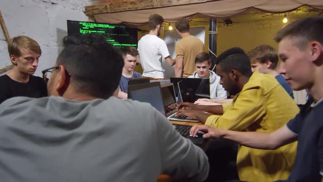Dolly shot of group of multiethnic young people sitting at desk together and writing codes on laptop computers, two of them standing at whiteboard and discussing something