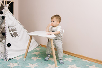 Cute baby sitting at the table and eating in the nursery white, gray and blue. Near the teepee and a bag of toys
