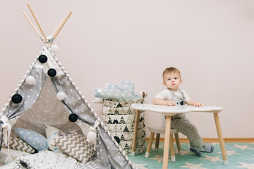 Cute baby sitting at the table in the children's room in white, gray and blue tones. Near the...