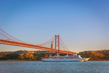 Lisboa, Portugal - May 15th 2018 - A medium size cruiser sailing through the Tejo River with the April 25th Bridge on the left side in a blue sky day