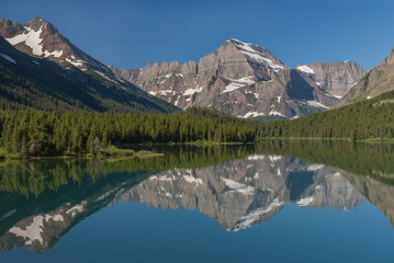 Reflections of the rugged mountains towering above the forested shores can be seen in Swiftcurrent Lake on a beautiful summer day in Glacier National Park