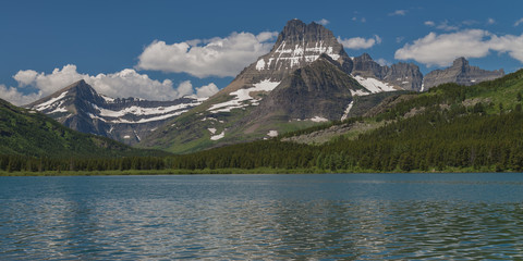 Rugged mountains with snow fields and small glaciers tower above the forested lake shore on a...