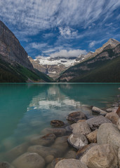 Rocks along the shore merge into a reflection of sky and mountains that lead to the mountains ...