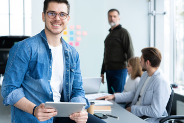 Young and creative. Handsome young man holding digital tablet and smiling while his colleagues discussing something in the background.