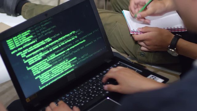 Close up shot of unrecognizable man using laptop computer with codes displayed on screen, someone sitting next to him and writing in notebook