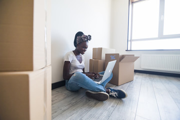Attractive African American woman unpacking boxes in her new home sitting on the floor with laptop...
