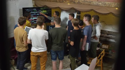High angle view of young man in glasses standing at whiteboard and explaining codes displayed on screen when delivering programming presentation at workshop