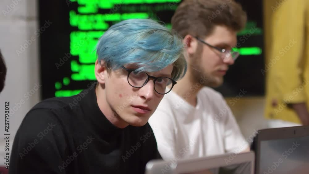Poster portrait of young man with blue hair wearing glasses while typing codes on laptop computer during pr