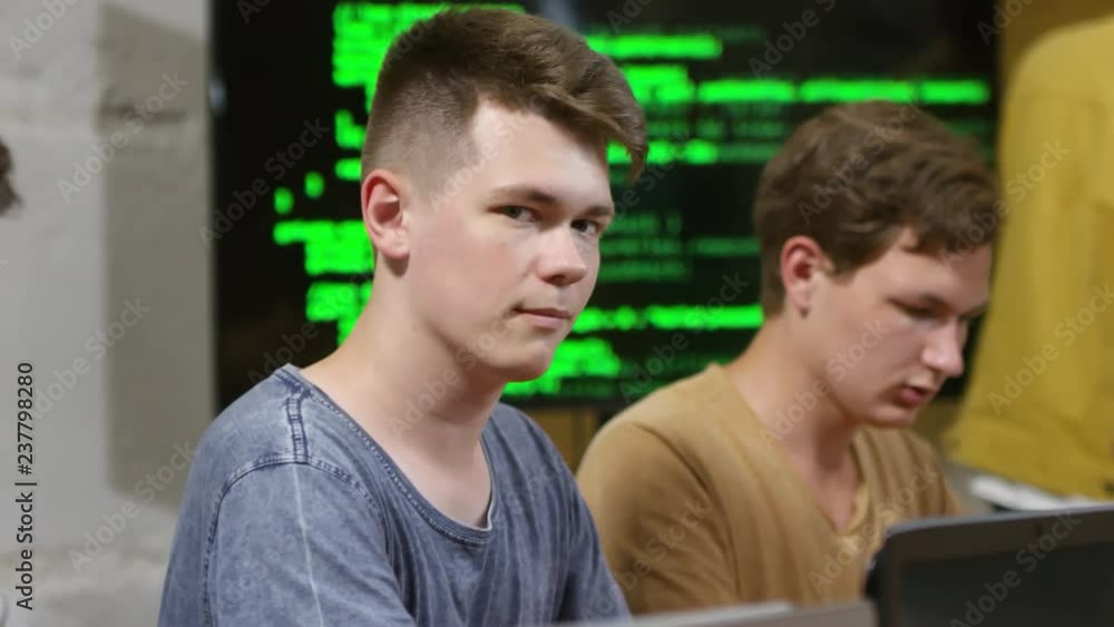 Poster portrait of young man sitting at desk and using laptop computer, big screen displaying codes behind 