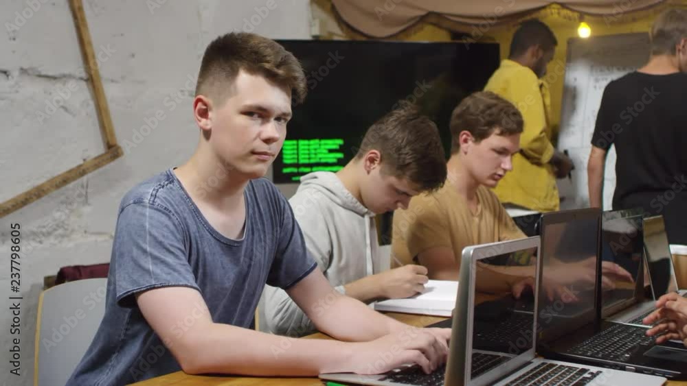 Poster portrait of young caucasian man sitting at desk during coding class, using laptop computer and looki