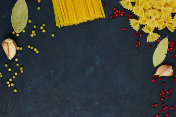 A variety of raw pasta next to the spices and spices on black concrete background. View from above. Copy space available. Italian food.