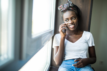 Cheerful afro american woman talking phone and sitting on windowsill at home