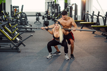 Beautiful girl doing squats under the supervision of the coach. Young couple is working out at gym. Attractive woman and handsome muscular man training together in modern gym.