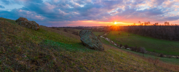 A skewed valley panoramic view of nature spring landscape. Spring landscape with big stone under the amber cloudy sunset. Valley with river
