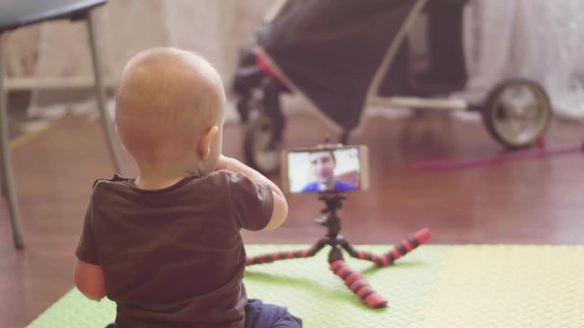 Father skyping on video call in a smartphone on tripod with his little son whos sitting on the floor. 4K slow mo back view