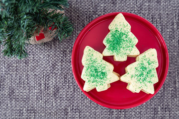 Three stacks of Christmas Tree shaped sugar cookies, with green sprinkles, on a red plate set out for Santa, and artificial tree on a brown background