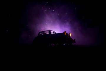 Fototapeta na wymiar Silhouette of old vintage car in dark foggy toned background with glowing lights in low light.