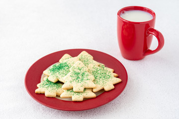 Christmas Tree shaped sugar cookies, with green sprinkles, on a red and white plate next to a red mug of milk set out for Santa, on a white background