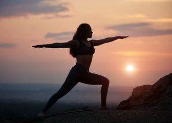Silhouette of slim long-haired girl with stretched arms in standing yoga pose outdoors on big rock...