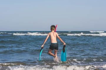 Fototapeta premium Happy teen boy in the swim flippers and snorkeling mask running оn the wave of the sea during summer vacation in the tropical resort town