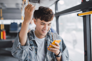 Young asian man is standing in the bus using phone and holding onto the bar while waiting to arrive...