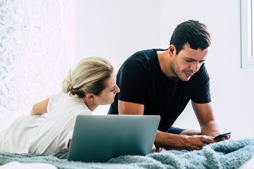 Young millennial modern couple at home in the bedroom morning time use technology devices like phone and laptop to check social media on internet - freelance everywhere work for alternative worker