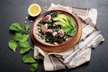 Pasta with cuttlefish ink (black noodles) with tuna pieces, spinach leaves, avocado slices, green peas, onions and sunflower seeds on a clay plate. An original pasta recipe. Useful and tasty dish