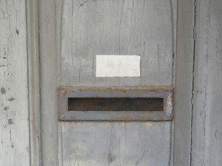 Torn and partially exfoliated layers of paint on an old wooden door with clear cracks, mailbox slot with rusted frame, white blank door plate without lettering