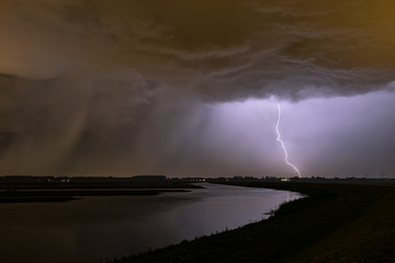 Omnious sky and powerful lightning strike with a rain shaft to the left near a lake in The Netherlands