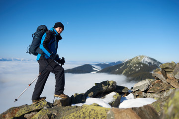 Tourist hiker with backpack and trekking poles climbing up on steep rocky mountain slope on background of blue sky, foggy valley filled with white clouds and snowy mountain tops.