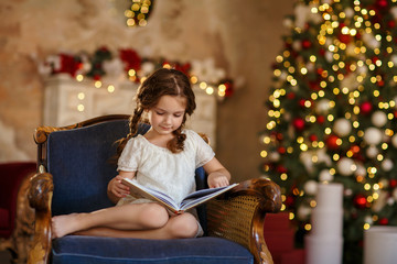 Little cute girl reads a book near christmas tree