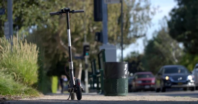 Electric rental scooter (without visible branding) parked on sidewalk next to street in Beverly Hills, Los Angeles, California. Day, rear view, focus on scooter with traffic approaching behind, 4K