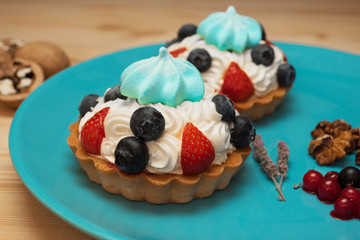 Two tartlets with white butter cream, blue marshmallow on top and strawberries and blueberries on blue plate and with walnuts and currant. The plate is on a wooden surface