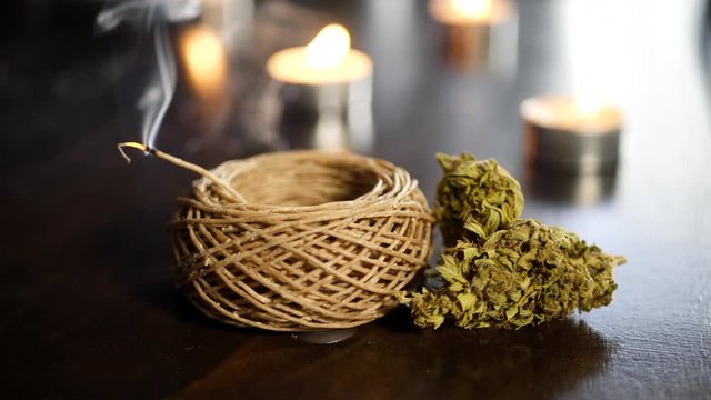 Marijuana buds for medicinal and recreational use next to a smoking hemp wick used to get high and bokeh candles on fire in the backgound.