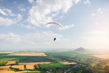 Professional paraglider in a cocoon suit flies high above the ground against the sky and fields with mountains