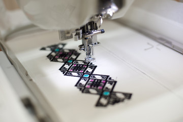 Close-up of sewing machine creating colorful abstract geometrical pattern on white fabric on blurred background. Modern technology, fashion and traditional clothing manufacturing concept.