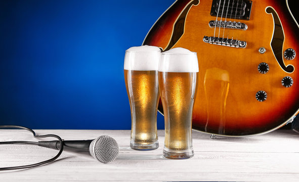 Two glass beer and microphone near electric jazz guitar on white wooden desk. Blue background. Free space for text