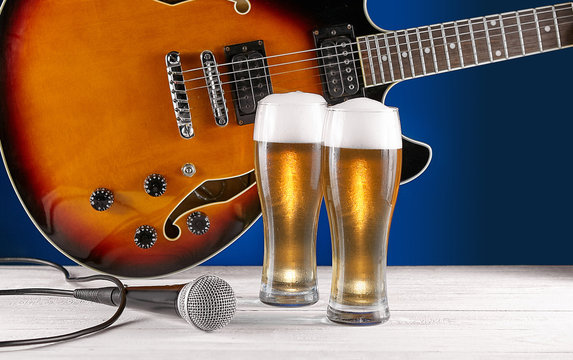 Two glass beer and microphone near electric jazz guitar on white wooden desk. Blue background