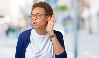 Young beautiful african american woman wearing glasses over isolated background smiling with hand over ear listening an hearing to rumor or gossip. Deafness concept.