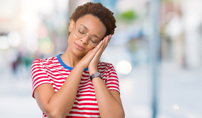 Beautiful young african american woman wearing glasses over isolated background sleeping tired dreaming and posing with hands together while smiling with closed eyes.